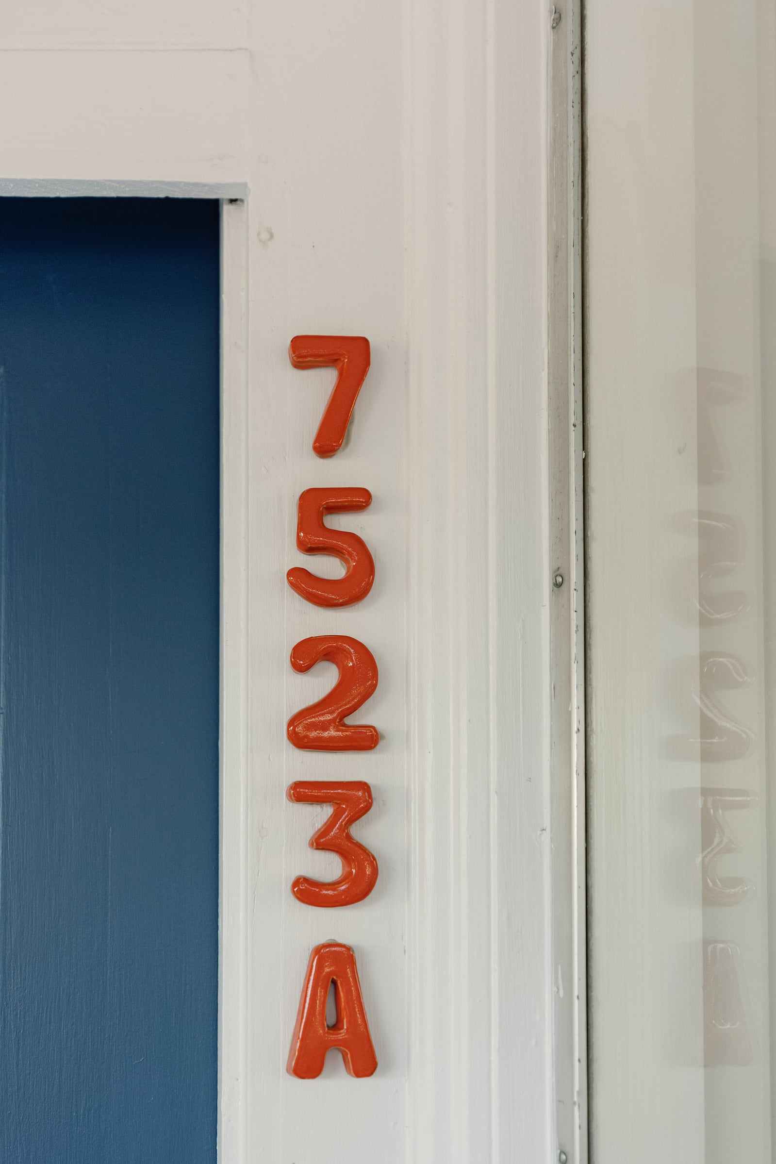 Glossy orange ceramic numbers reading 7523A, stacked down the white door frame beside the blue door, with their reflection in the front window.