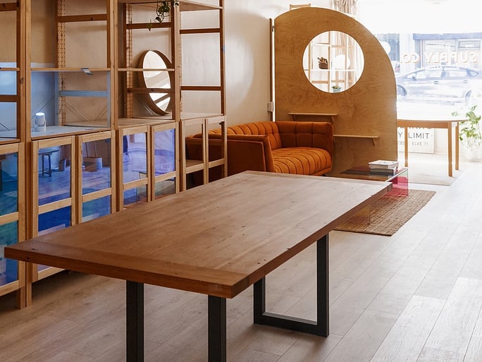 The big table's long honey-colored top of reclaimed wood flooring in the foreground, with the pine shelving, the orange sofa, the arched pony wall and the sunlit front window behind it.