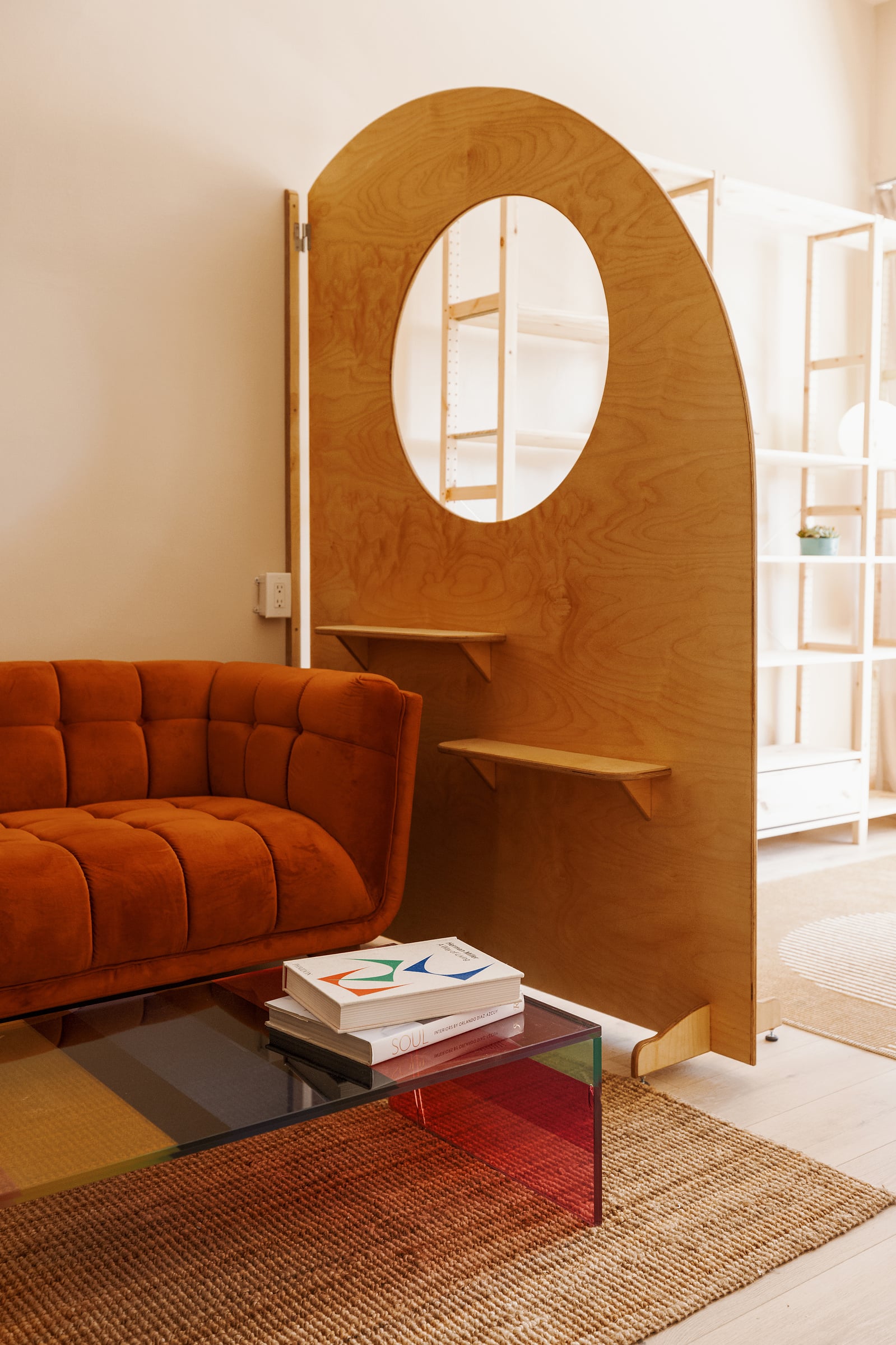 The arched plywood pony wall, with a round window cut through it and two small shelves, beside the rust velvet sofa and the rainbow acrylic coffee table.