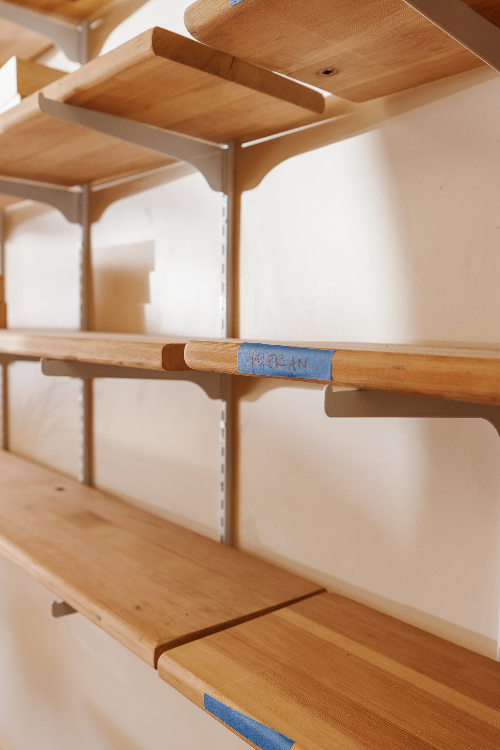 Reclaimed bleacher-seat shelves with rounded edges on white wall brackets, one labeled with blue painter's tape reading Kieran.