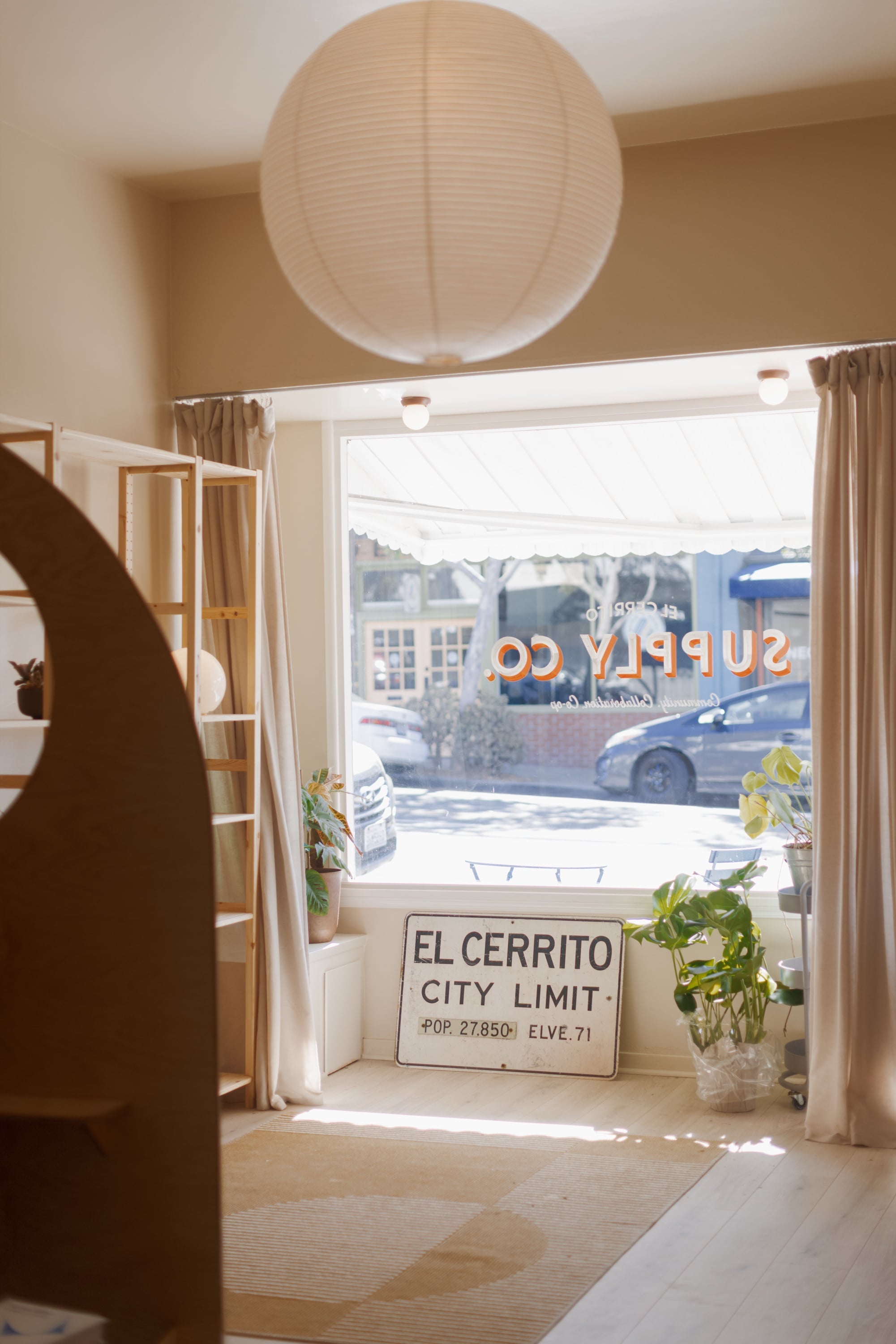 Inside the shop facing the sunny front window, under a paper lantern, with the Supply Co. lettering on the glass and an old El Cerrito city limit sign propped on the floor below it