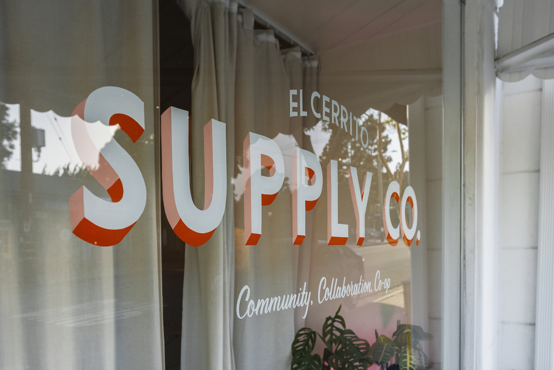 The finished hand-painted window sign: El Cerrito Supply Co. in white block letters with an orange and pink drop shade, over the script line Community, Collaboration, Co-op.
